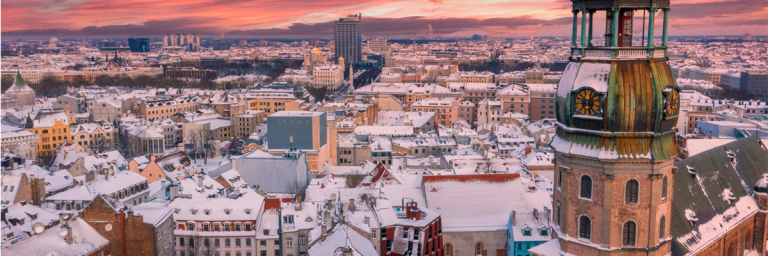 Vista aérea de la Ciudad Vieja de Riga, Letonia, cubierta de nieve. El cielo al atardecer presenta tonos anaranjados y púrpuras, destacando la torre de ladrillo de la iglesia de San Pedro en primer plano.