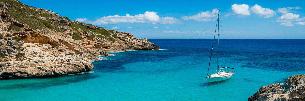 Grupo de viajeros singles disfrutando a bordo de un velero en aguas turquesas de Mallorca, con acantilados y vegetación mediterránea de fondo al atardecer. Reflejo del sol en el agua.